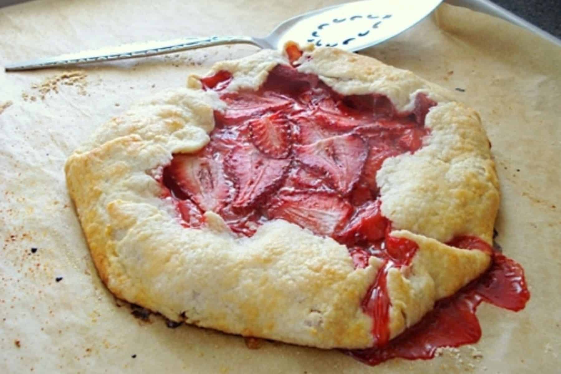 Horizontal image of strawberry crostata on baking sheet with serving piece behind it.