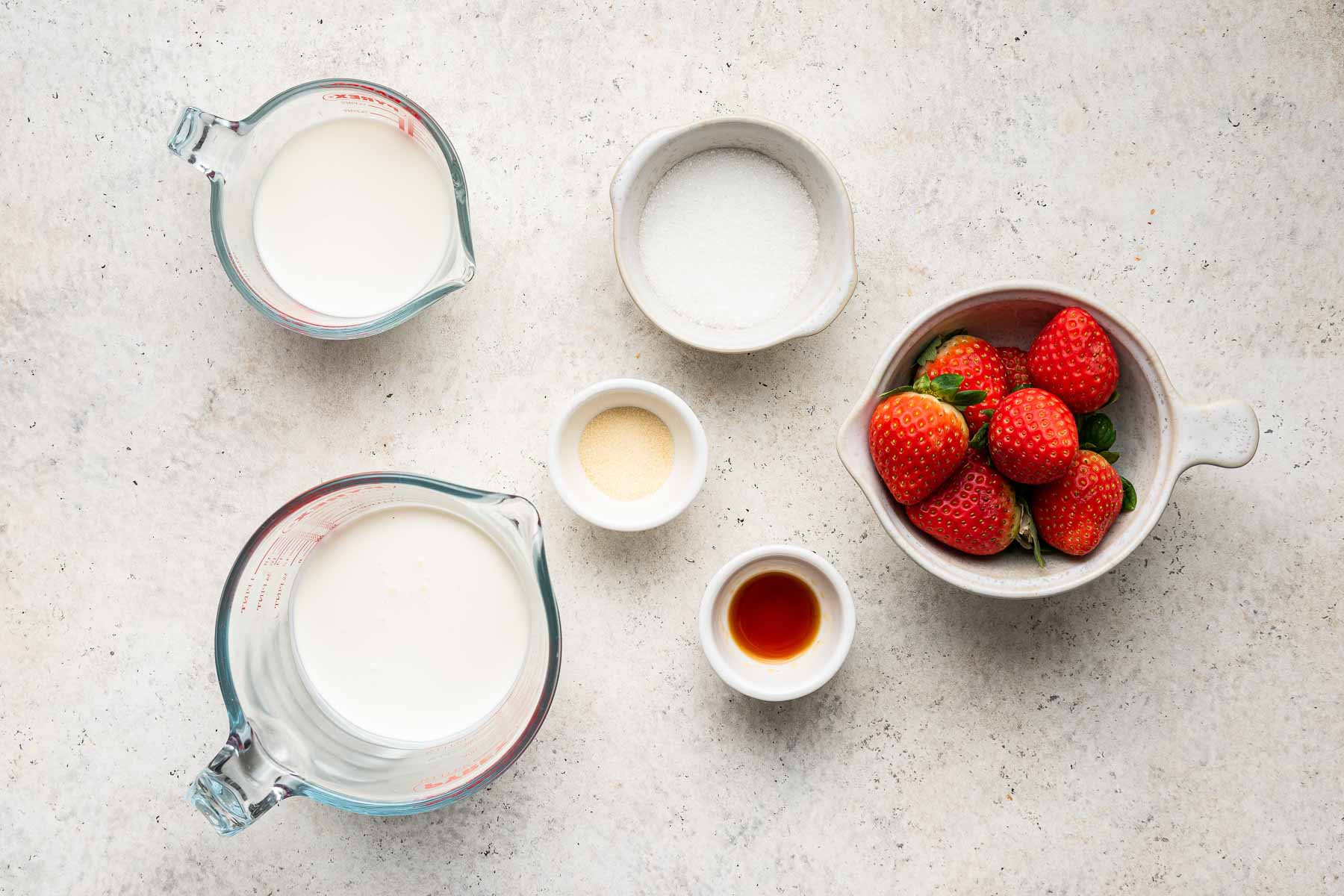 Grey background with milk, cream, sugar, vanilla, gelatin and strawberries in small bowls.