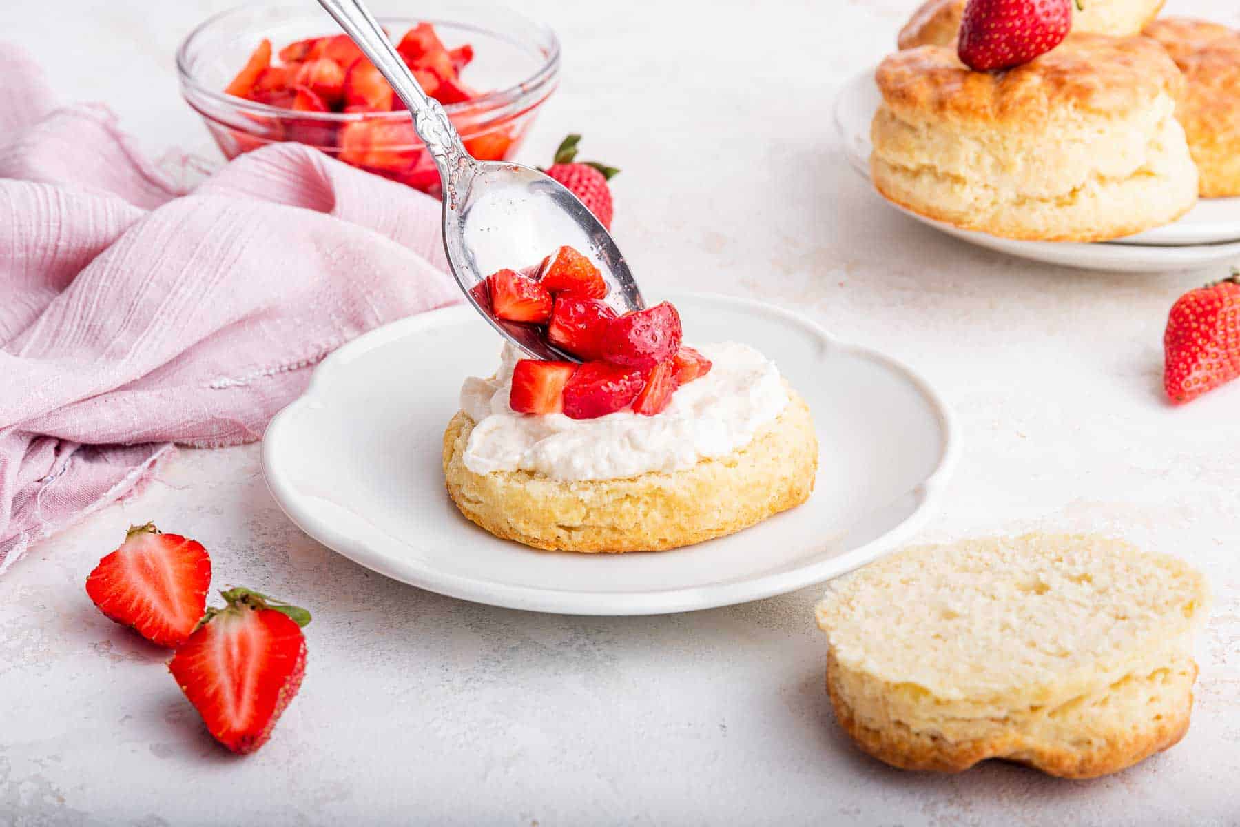 Horizontal image of biscuit cut in half, being layered with whipped cream and strawberries.