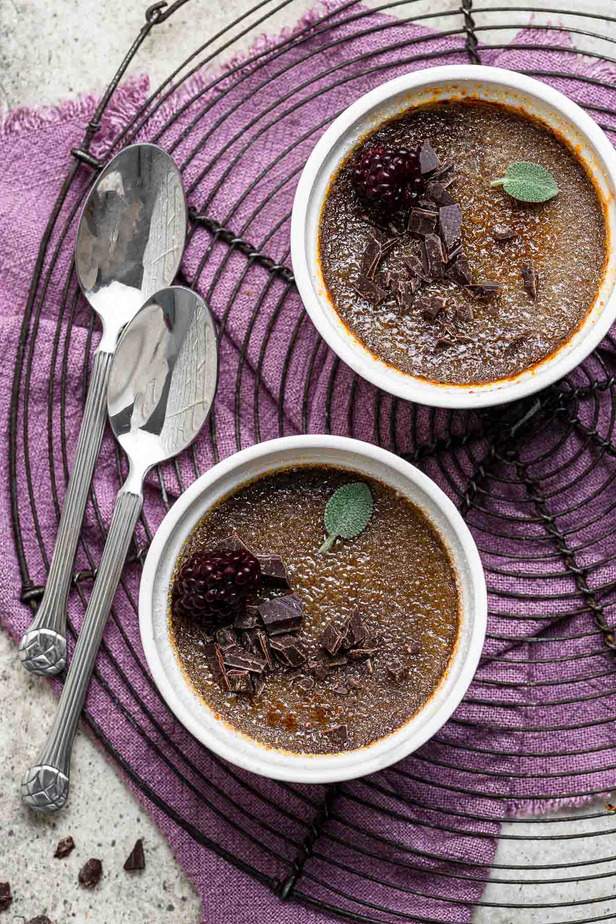 Two ramekins of chocolate creme brulee on wire rack with spoons.