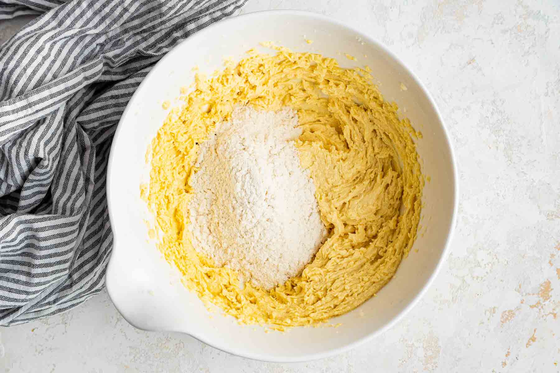 Half of white flour on top of yellow batter in white bowl with kitchen towel on side.