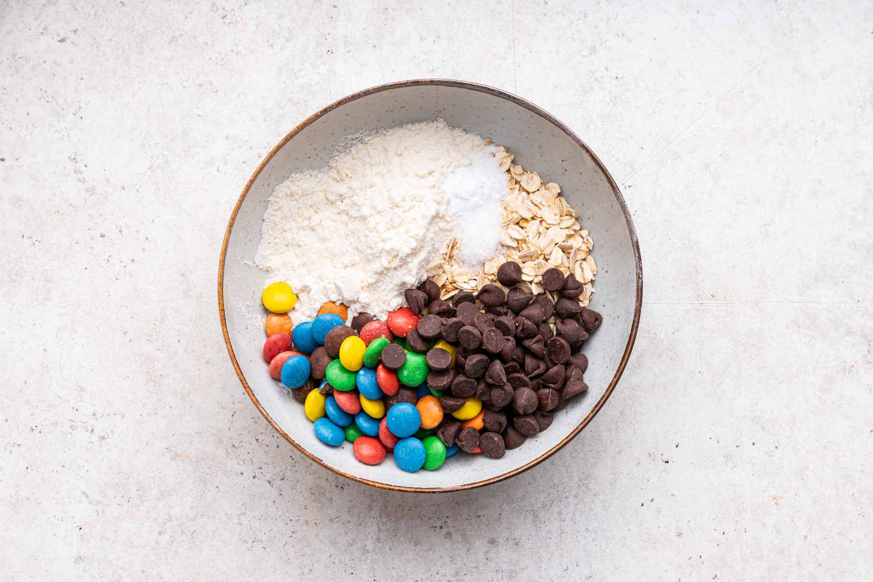 White bowl with flour, chocolate chips, oats and colorful chocolate candies.