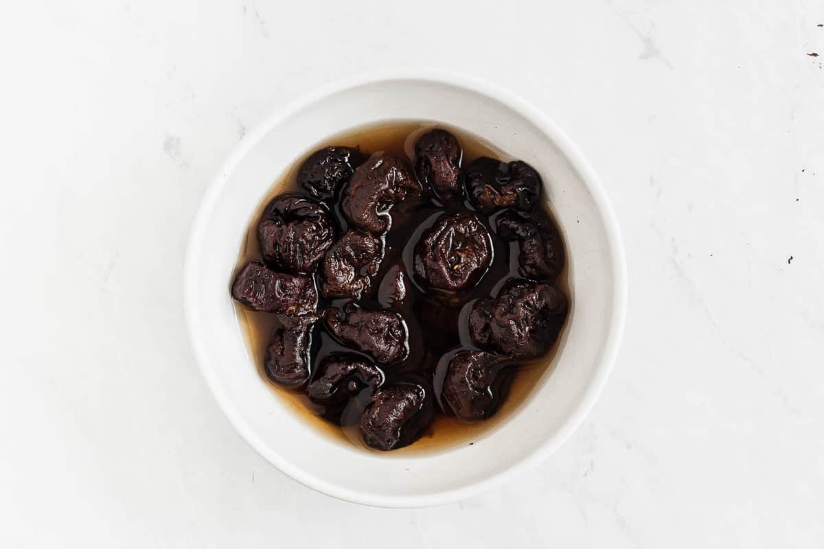Prunes soaking in hot water in a white bowl.