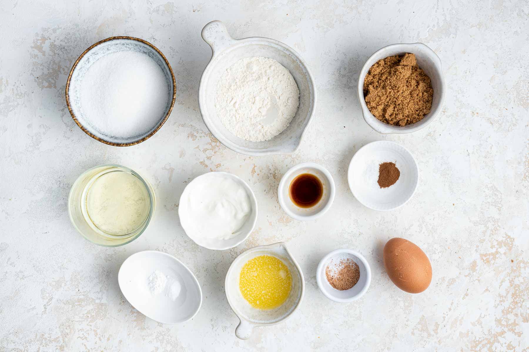 Small bowls of flour, sugar, brown sugar, egg, and vanilla on white surface.