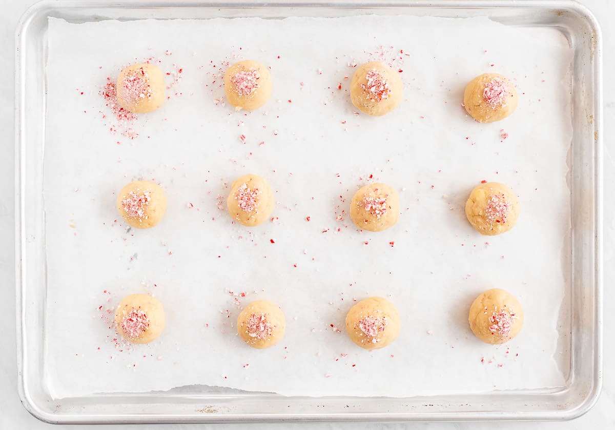 Cookie sheet with raw candy cane sugar cookie dough in balls with extra crushed peppermints on top.