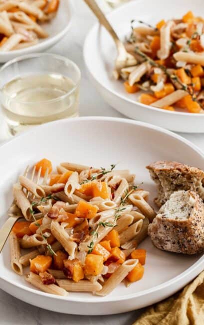 Two bowl of pasta with butternut squash cubes and bread on the side.