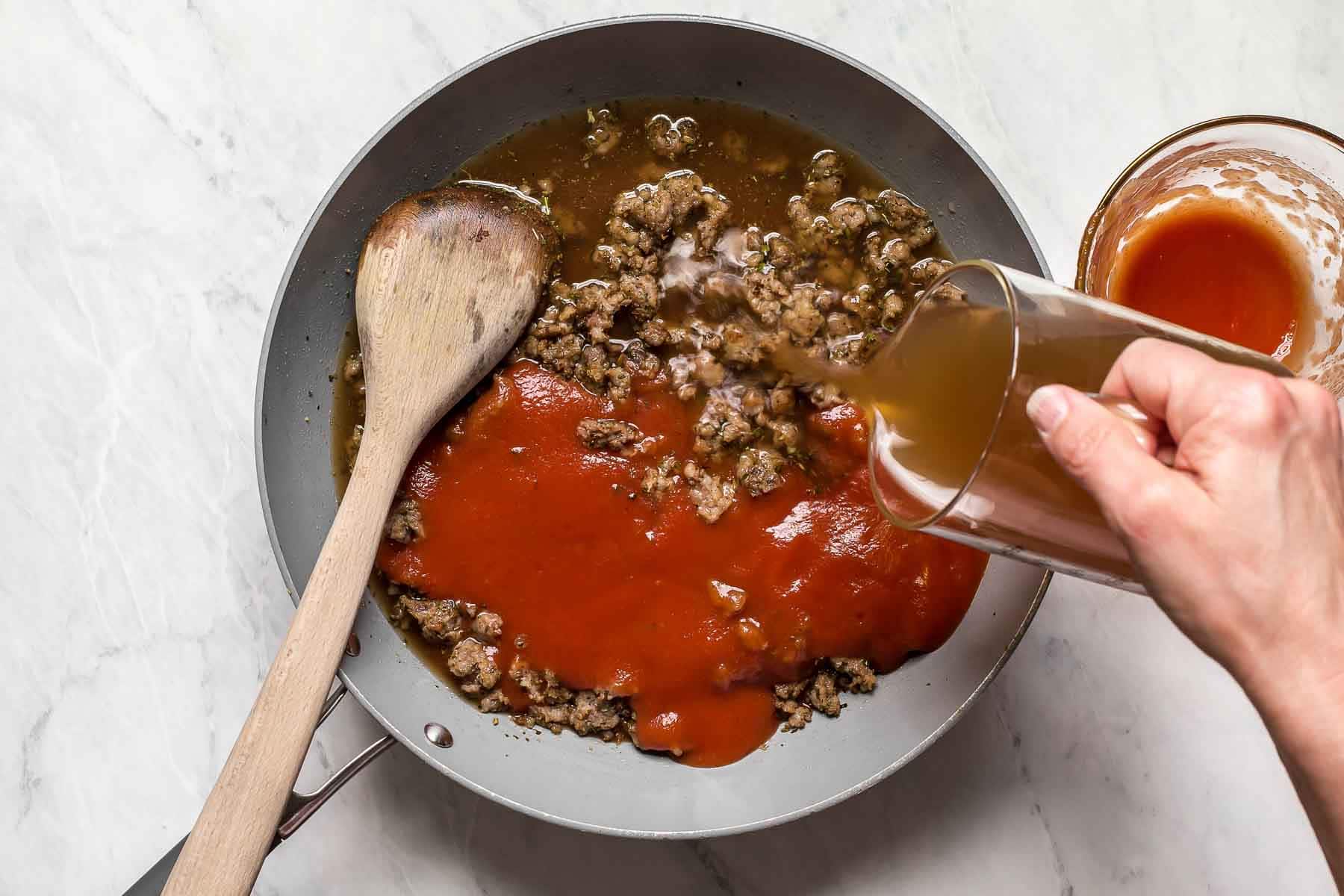 Tomato sauce pouring into skillet with ground sausage.