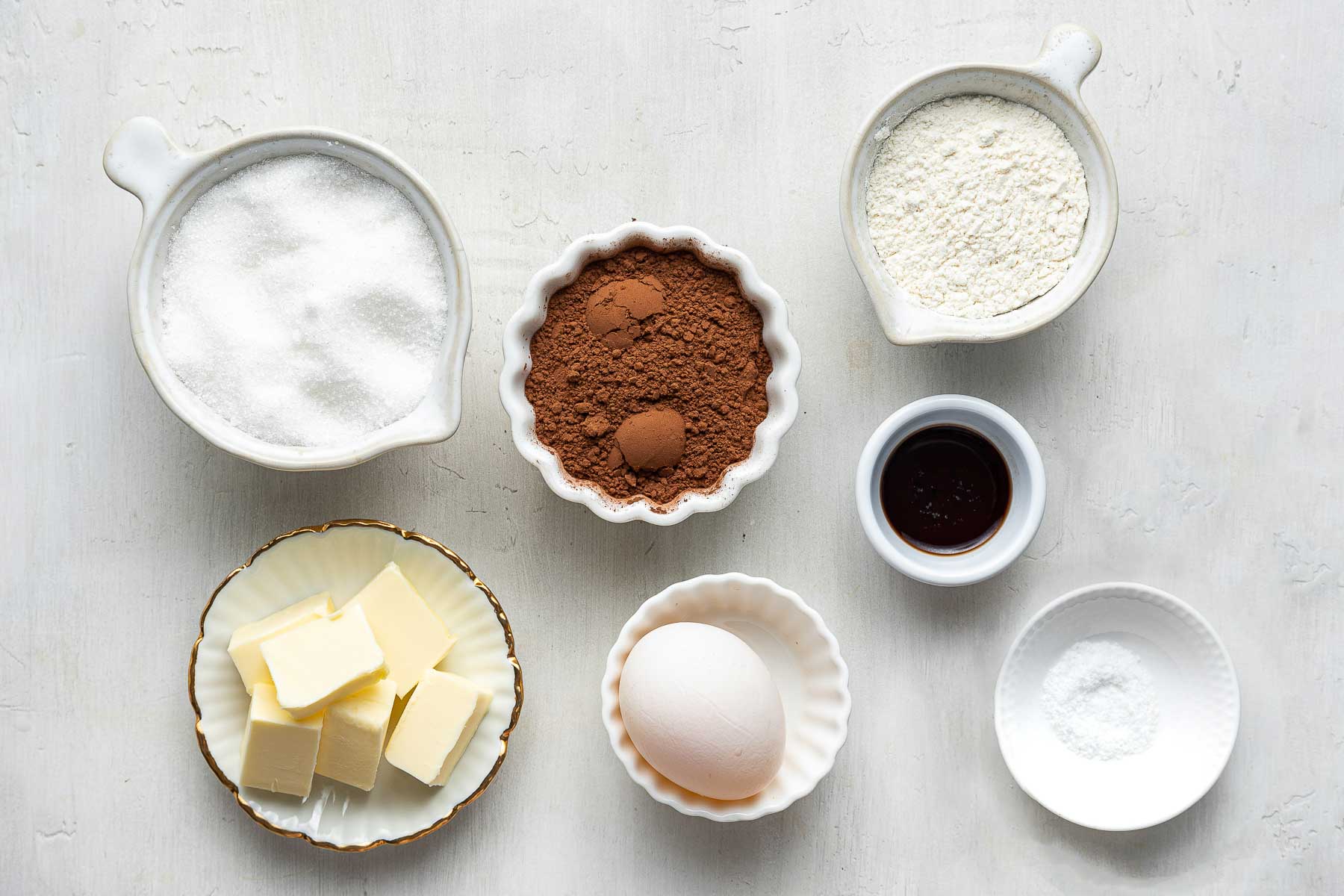Sugar, butter, flour, egg and cocoa powder in small bowls on white surface.
