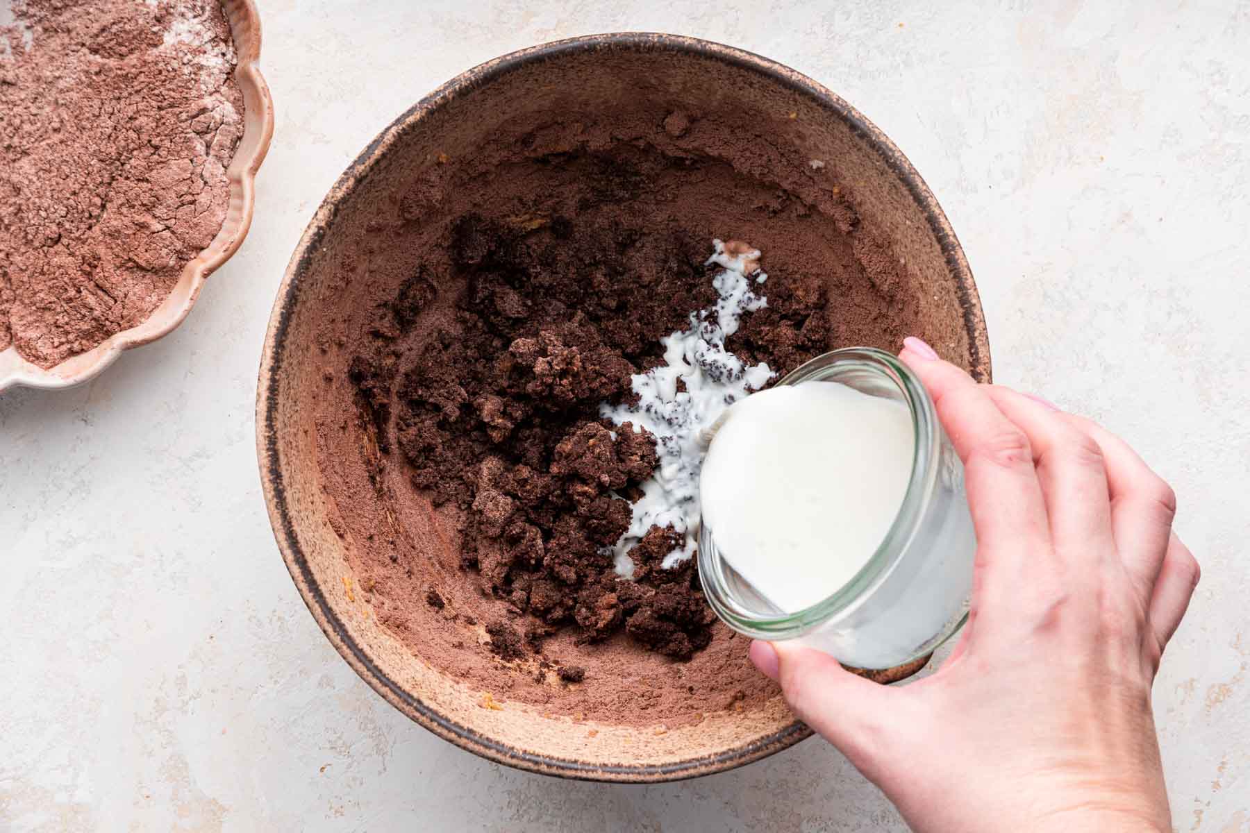 Hand pouring milk into brown dough in brown bowl.