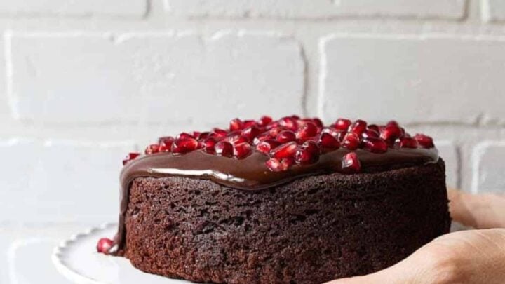 A mini chocolate cake with pomegranate ariels on top on a white platter held with one hand.