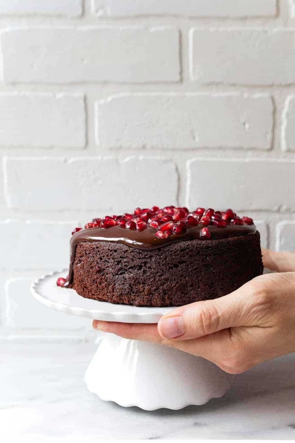 A mini chocolate cake with pomegranate ariels on top on a white platter held with one hand.