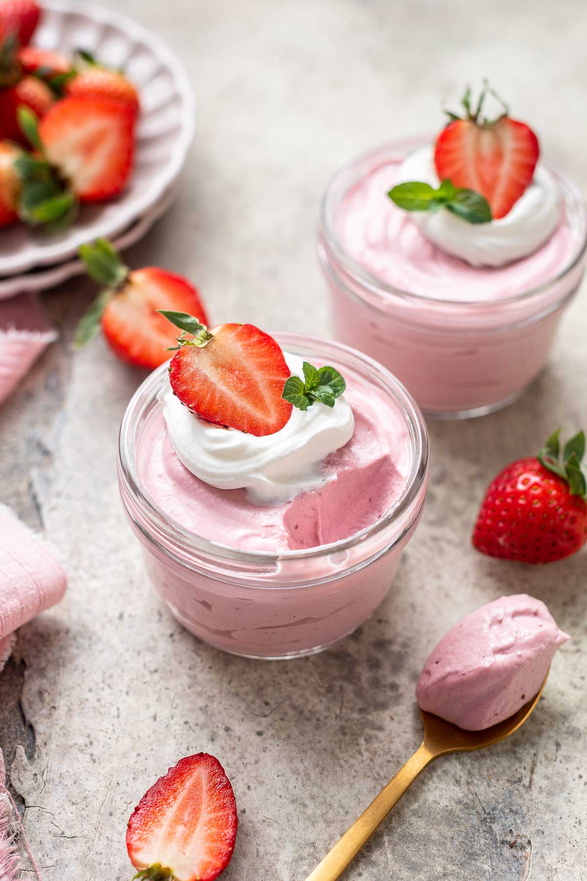 Two glass jars of strawberry mousse garnished with whipped cream and fresh strawberries.