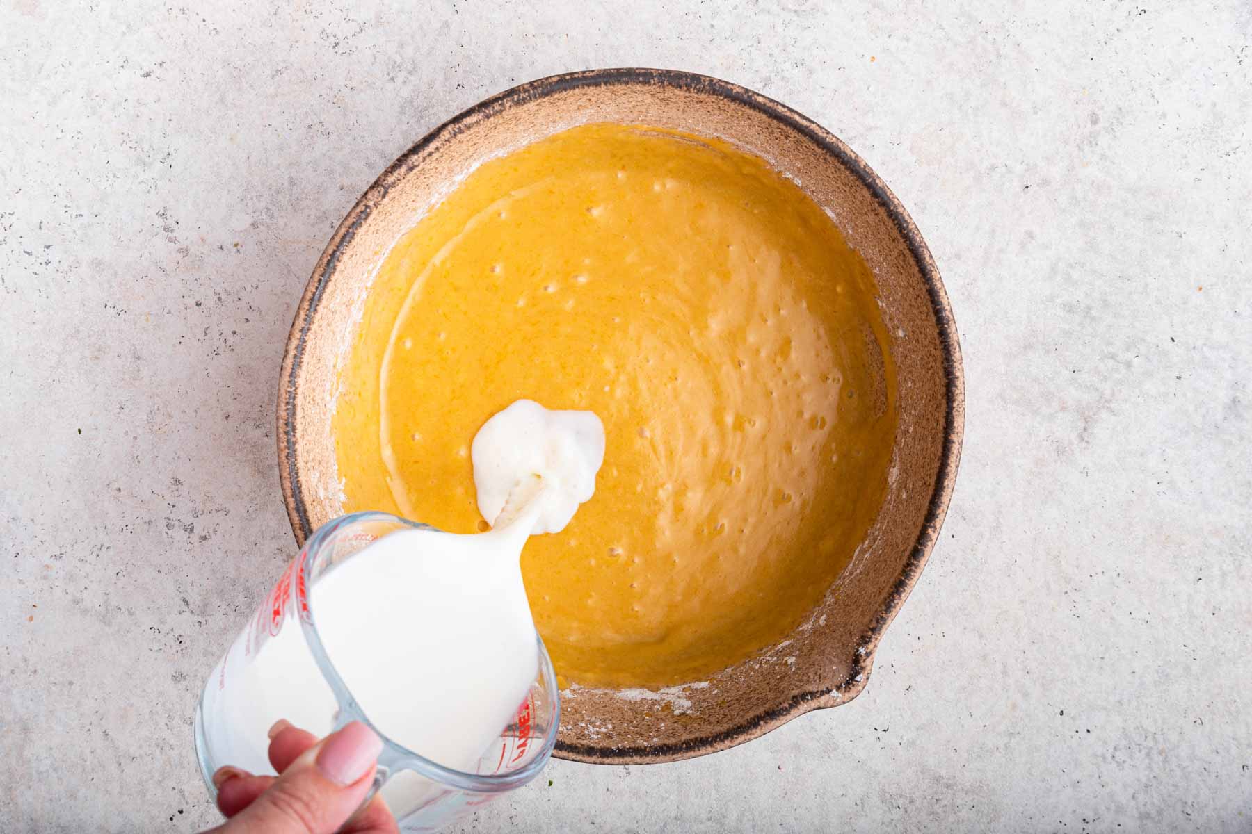Pouring milk into yellow cake batter in brown bowl.