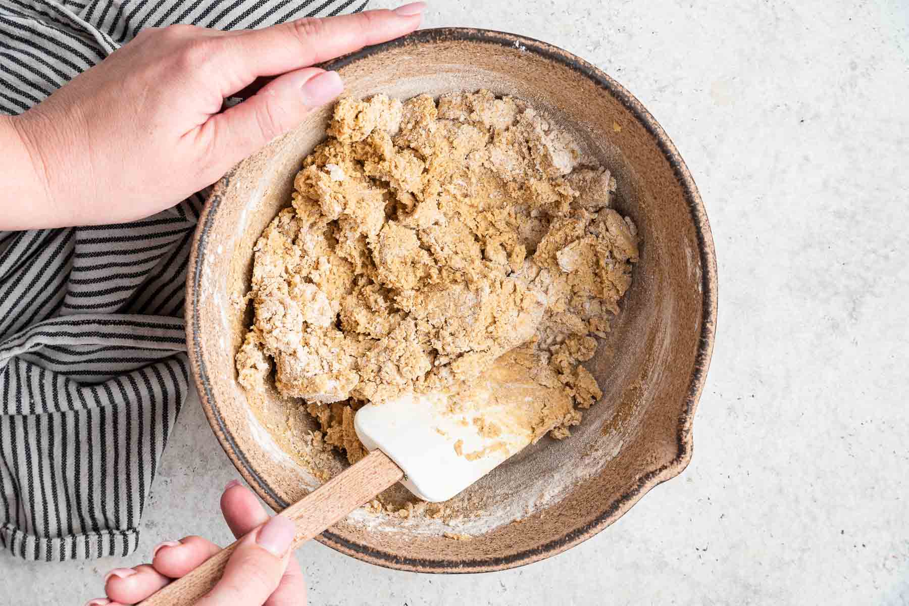 Two hands stirring dough together in brown bowl with white spatula.