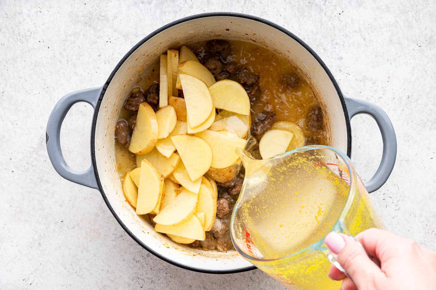 Sliced potatoes in a dutch oven with chicken broth being poured on top.
