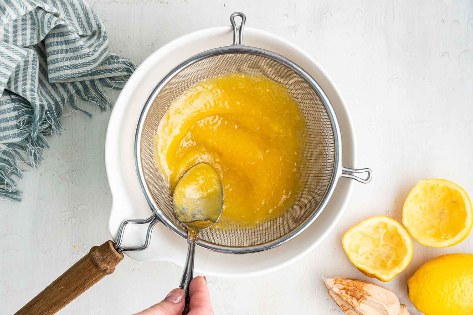 Spoon pushing yellow batter through strainer over white bowl.