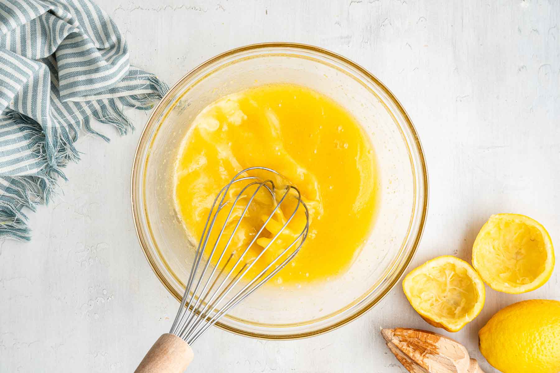 Whisk in a bowl with yellow liquid and lemon peels on side.