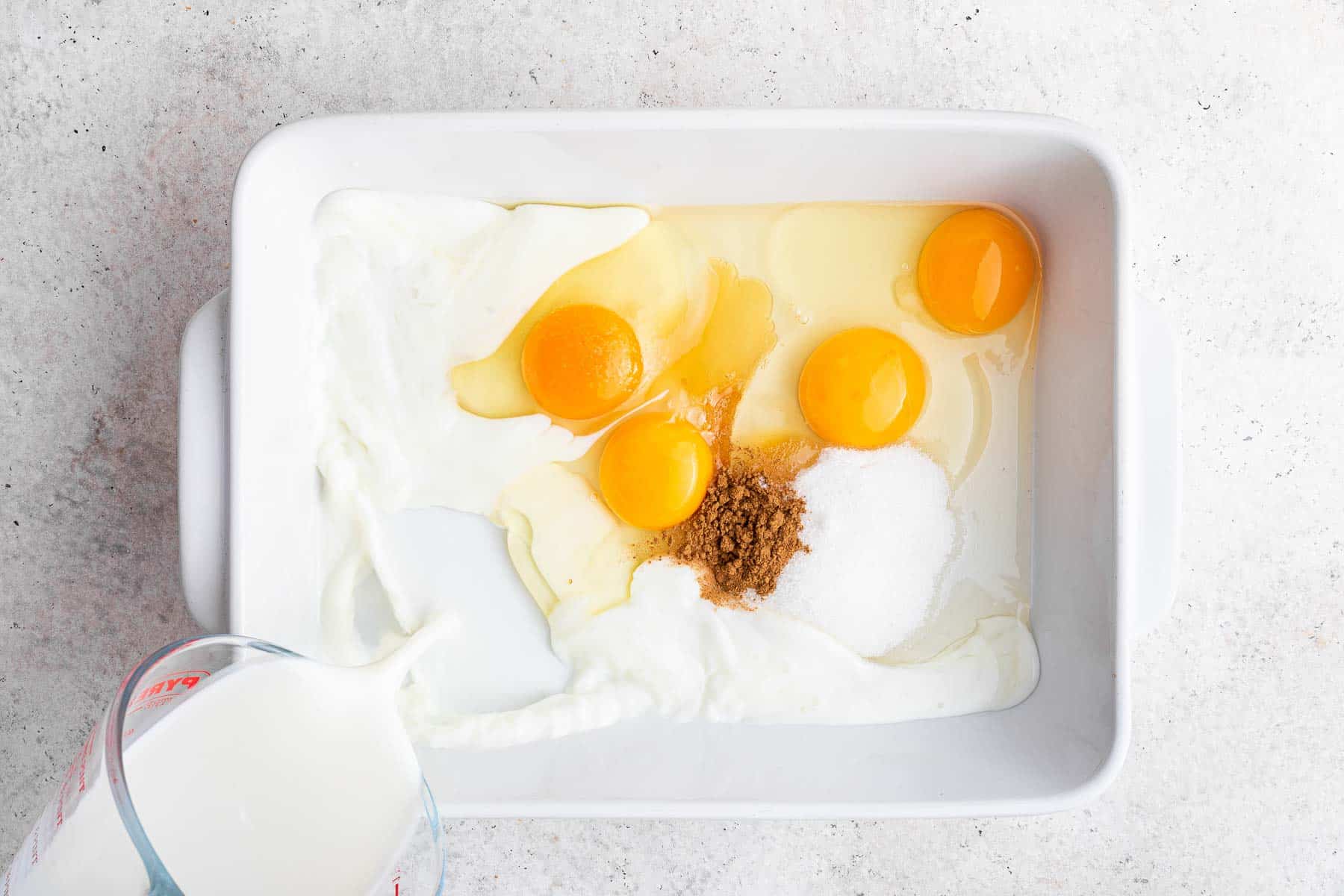 White casserole dish with eggs, cinnamon and milk being poured in.