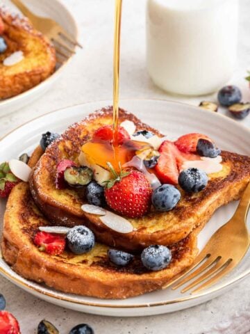 Brioche French toast for two on plate with fresh berries and syrup being poured on top.