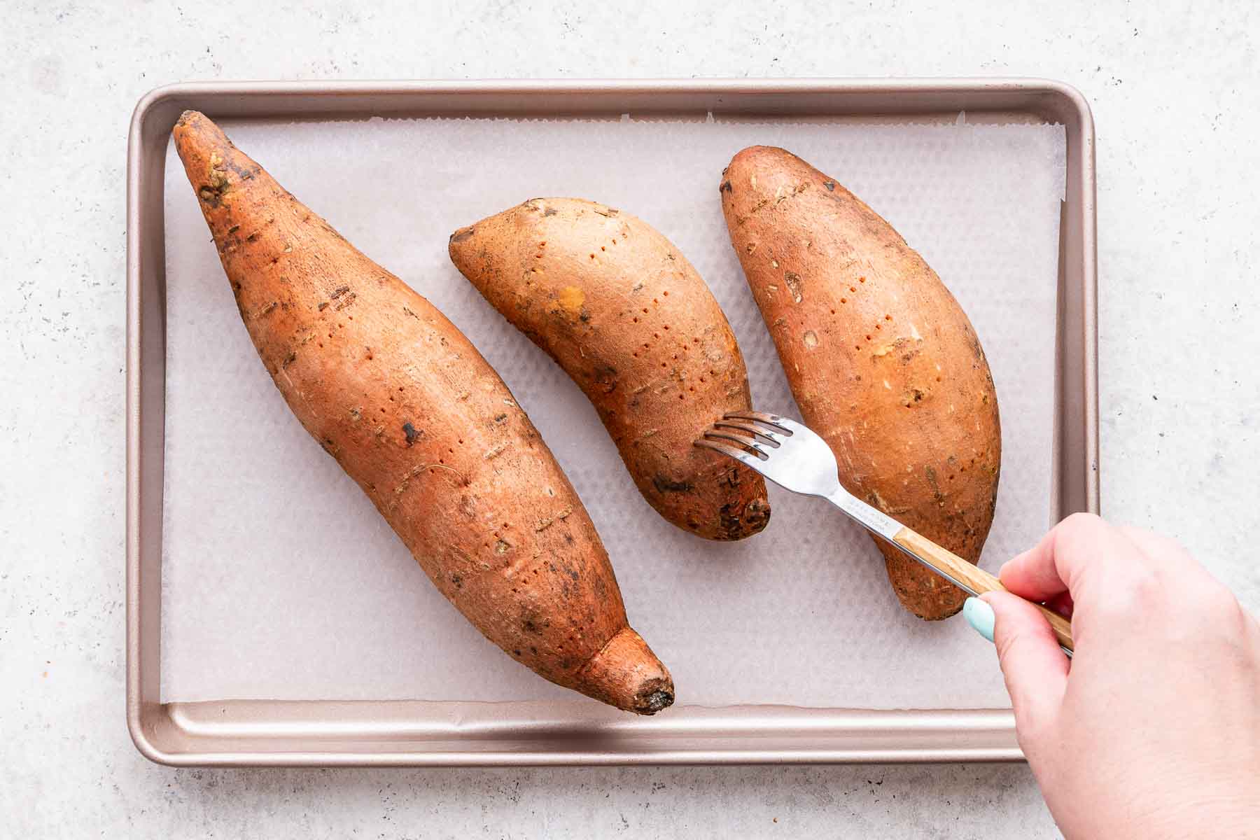 Hand pricking a fork into raw sweet potatoes on a baking dish.