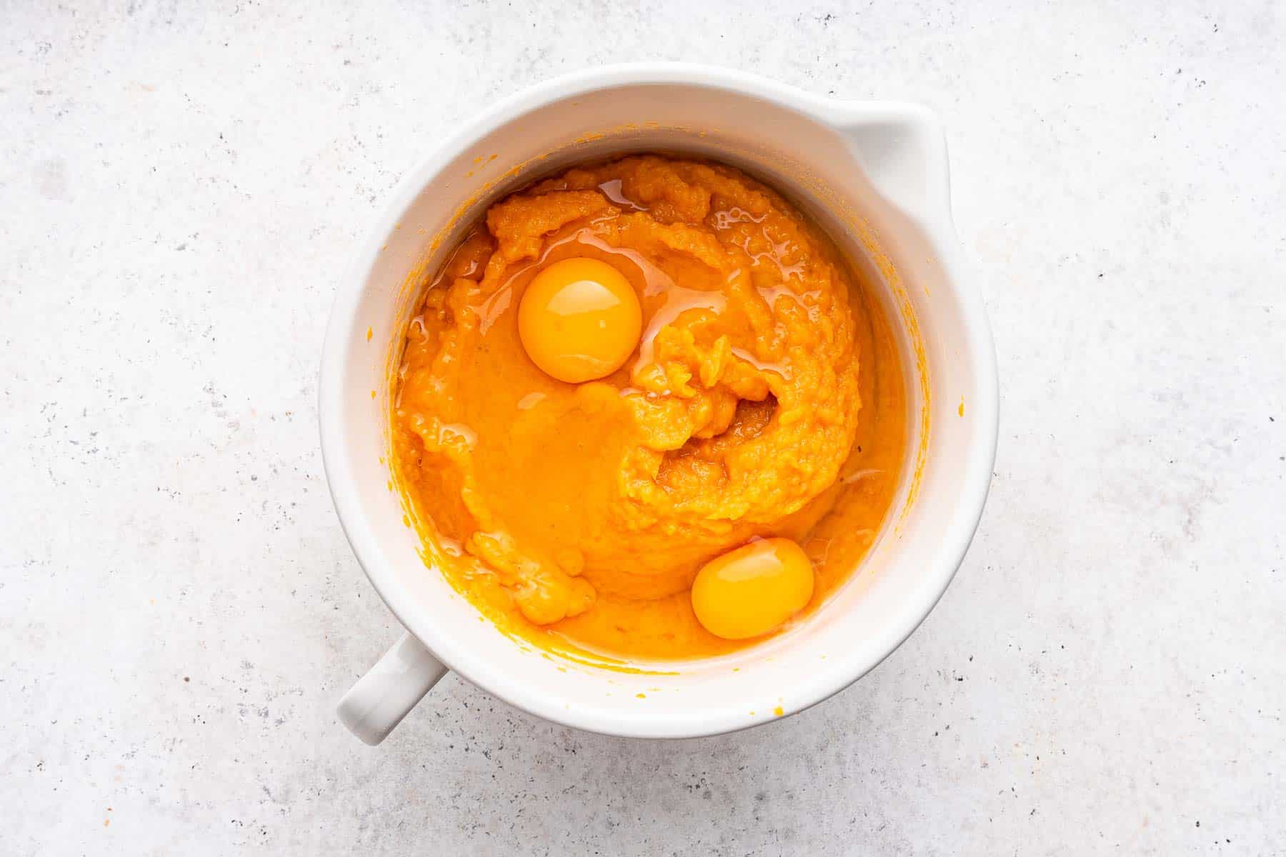 Adding whole eggs to orange batter in white round bowl.