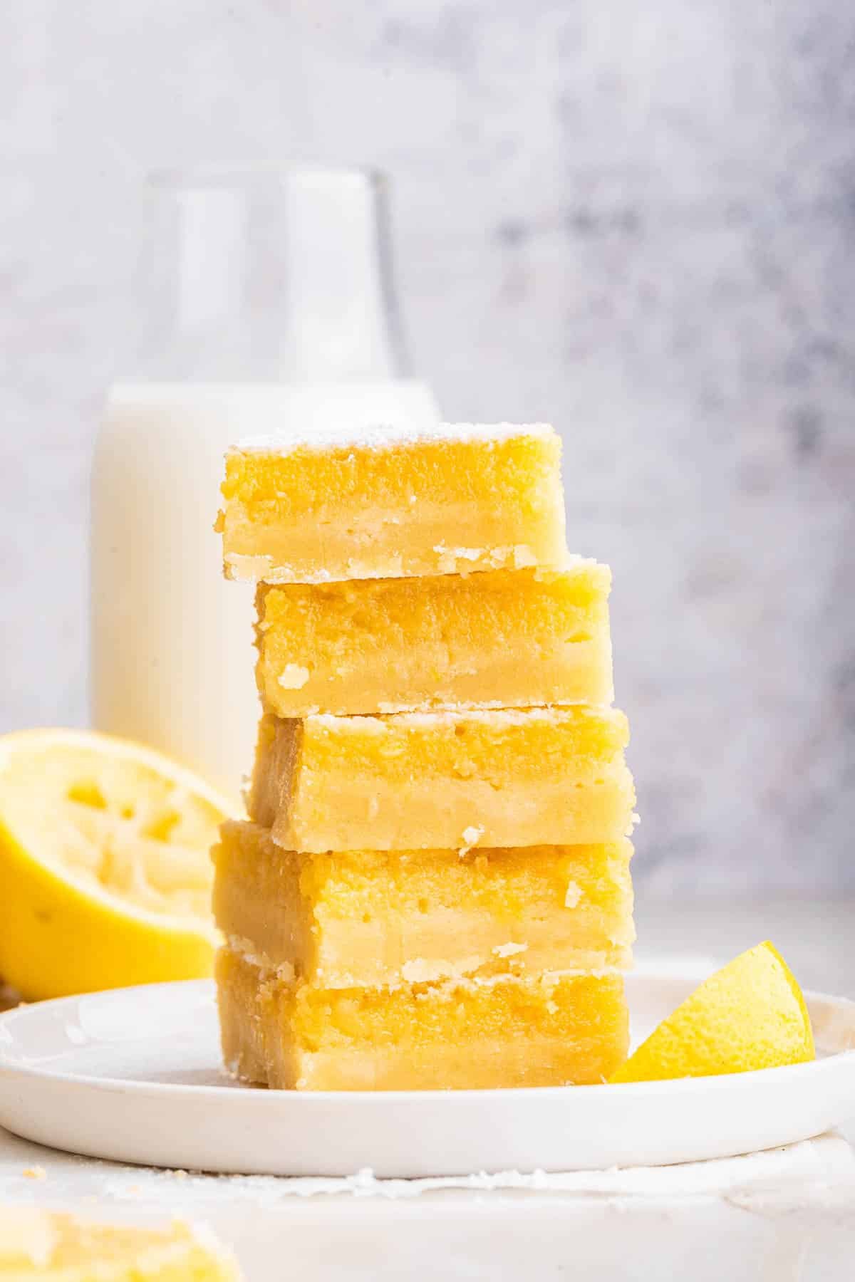 A stack of five lemon bars on a white plate with milk in the background.