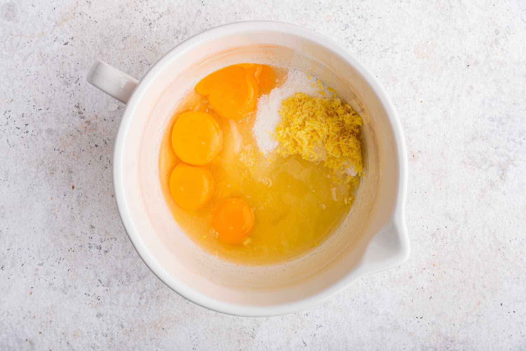 White bowl with eggs, zest, sugar and juice inside.
