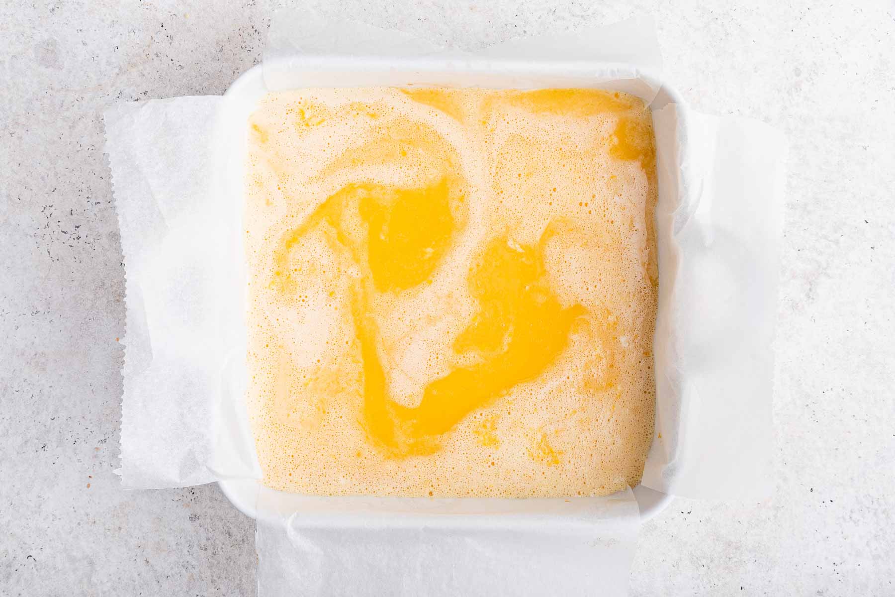 Yellow wet batter being poured in a white square pan lined with parchment paper.