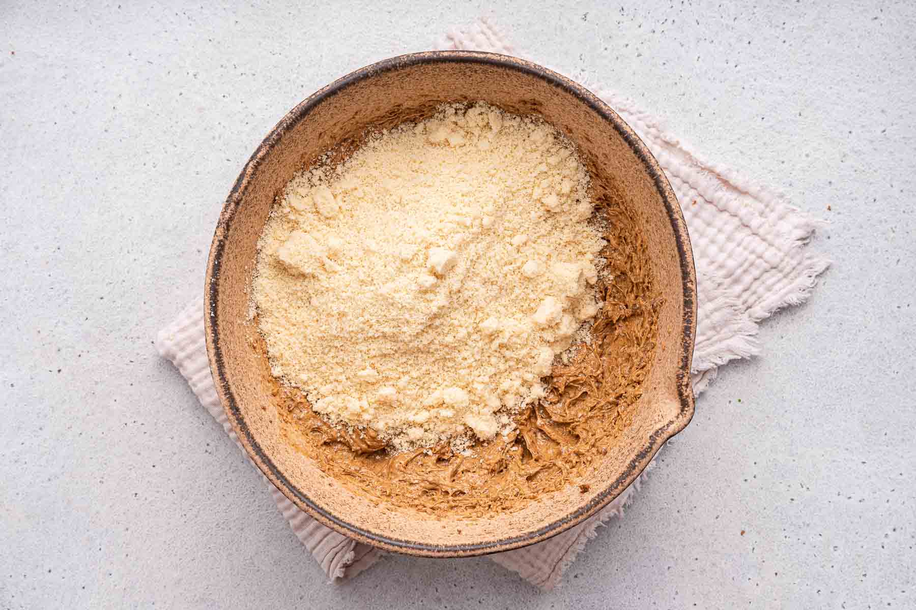 Adding yellow flour on top of raw brown cookie dough in mixing bowl.