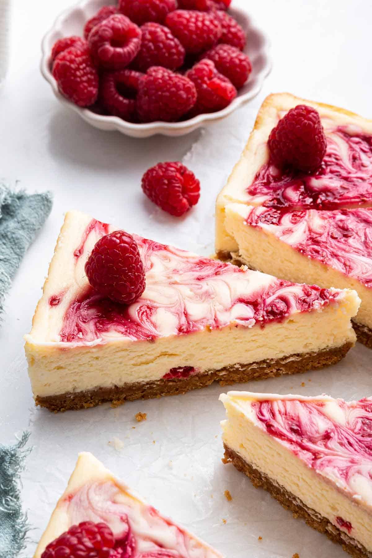 White chocolate raspberry cheesecake with swirls and a bowl of berries behind a slice.