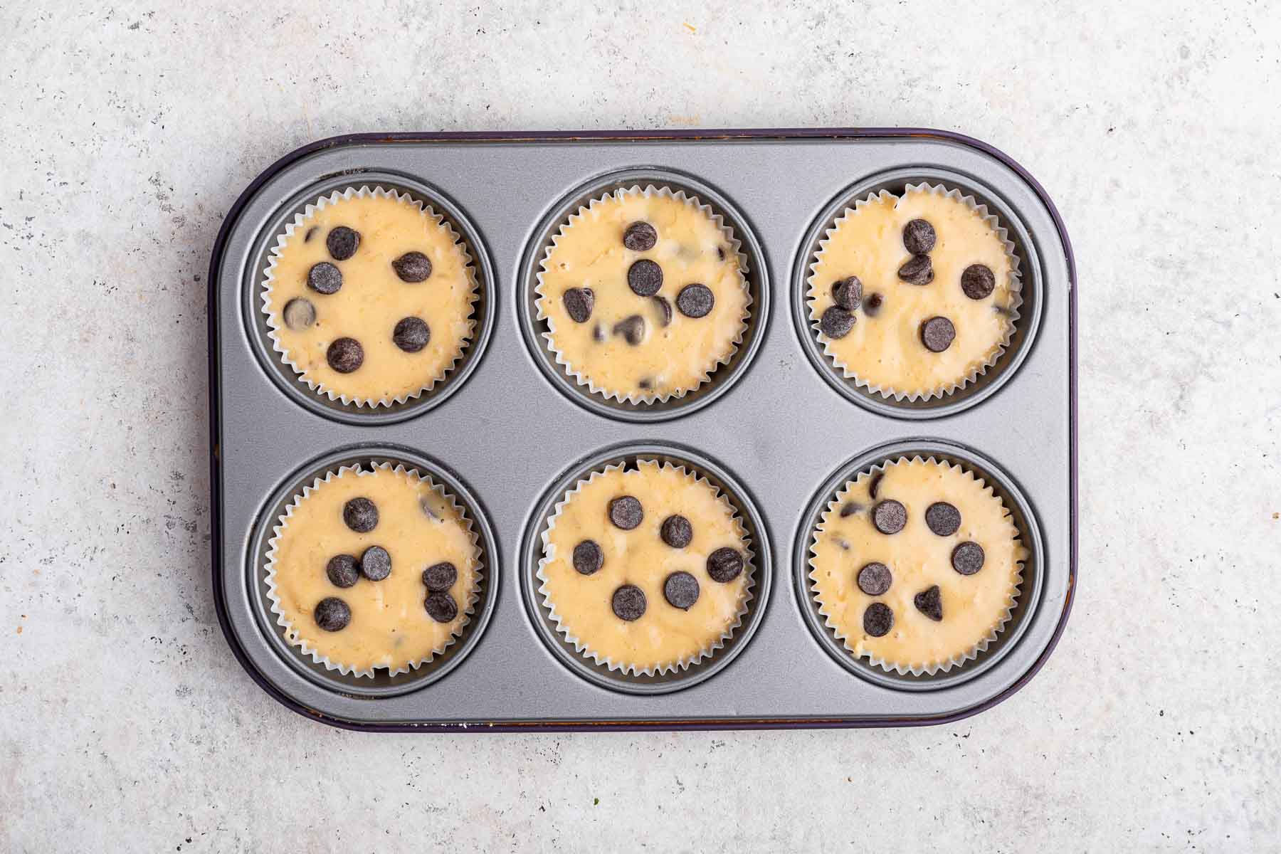Six unbaked yellow muffins with chocolate chips sprinkled on top.