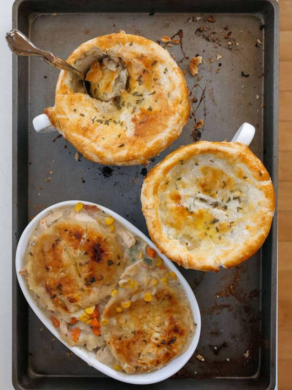 Two white soup mugs and a lomg oval baking dish with a browned top crust of chicken pot pie on a grey baking sheet.