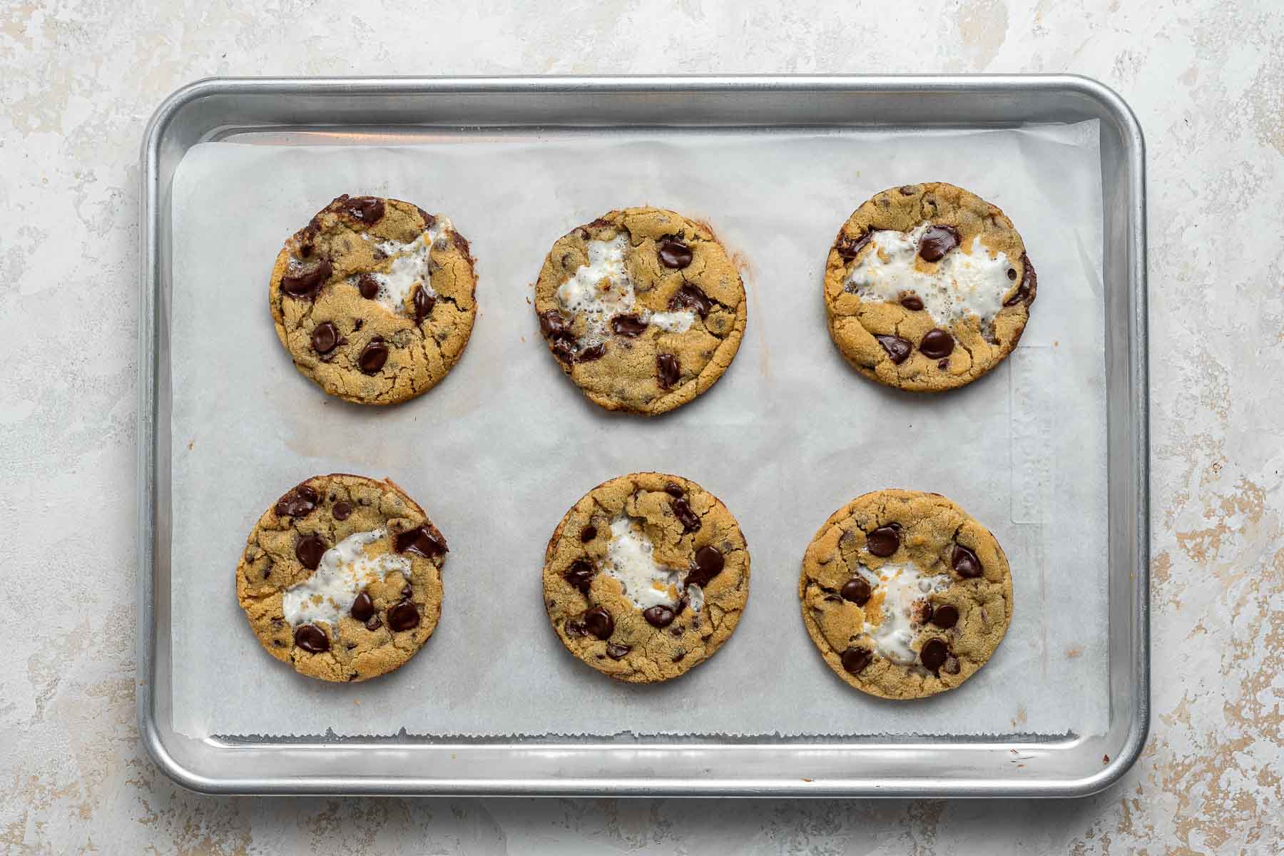 Six chocolate chip cookies with marshmallow centers on a cookie sheet.