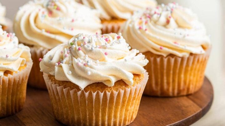 Wooden plate of decorated white cupcakes with sprinkles.