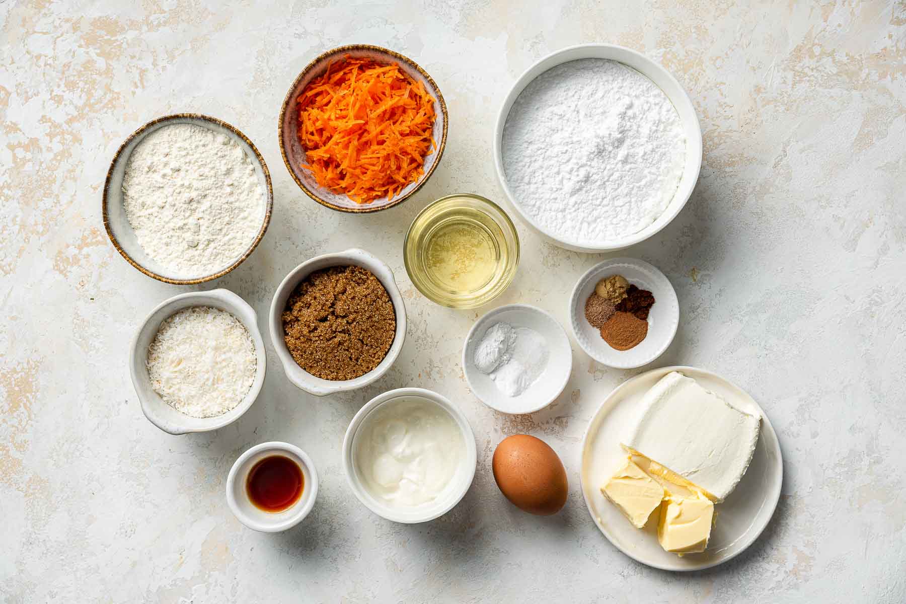 Ingredients for carrot cake in small bowls on white table.