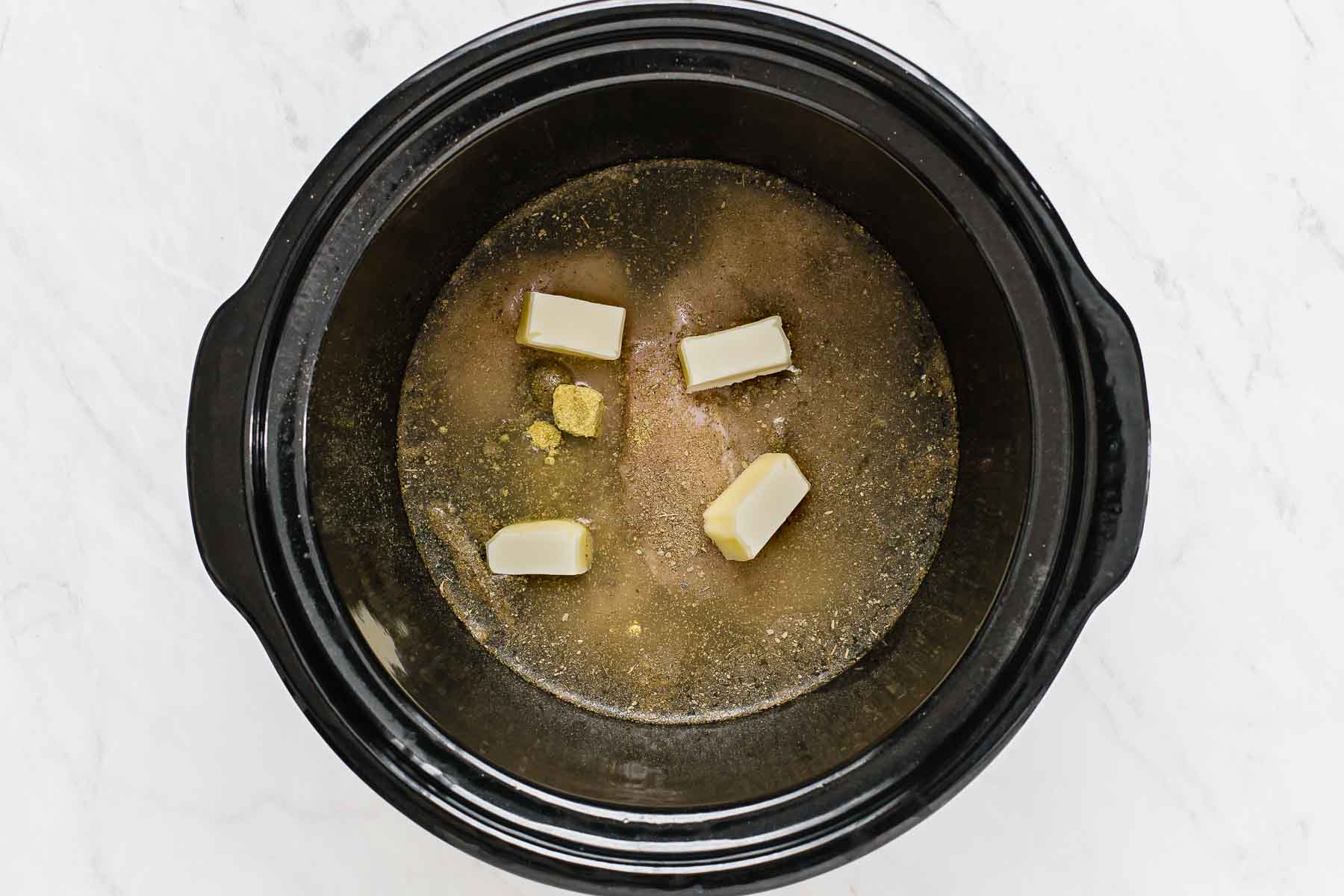 Chicken breasts with liquid and butter slices on top in crockpot.