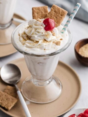 White cheesecake milkshake in glass with whipped cream, a cherry and graham crackers.