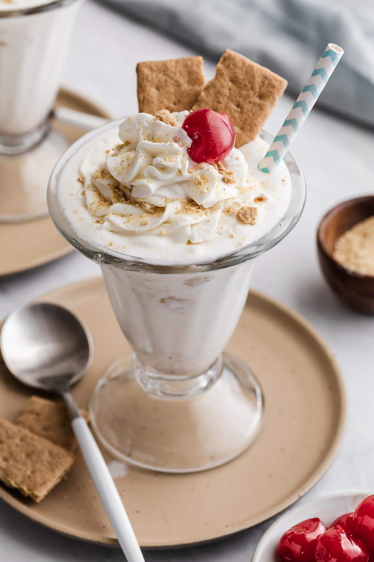 White cheesecake milkshake in glass with whipped cream, a cherry and graham crackers.