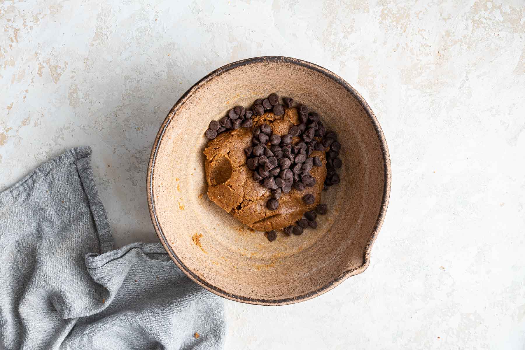 Brown dough in a brown bowl with a scoop of chocolate chips on top.