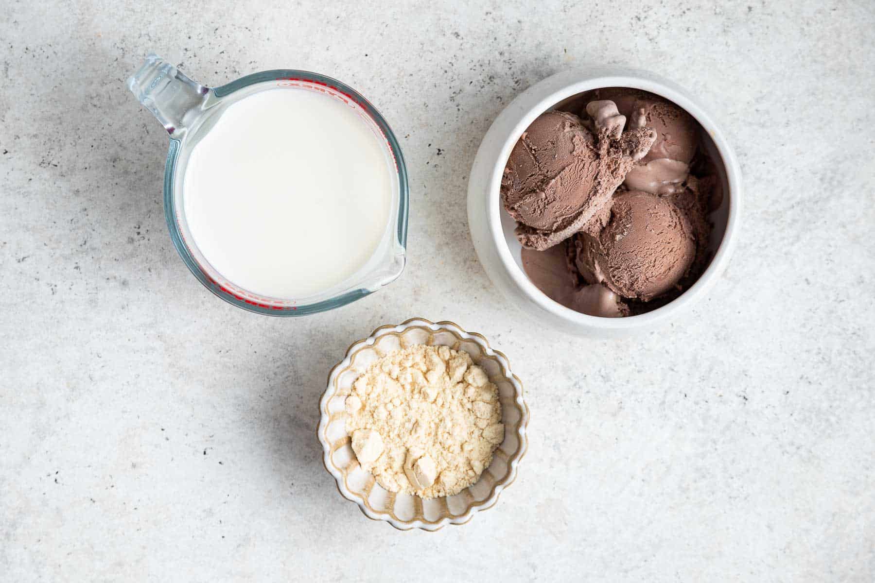 Three bowls with chocolate ice cream, milk, and milk powder on grey counter.