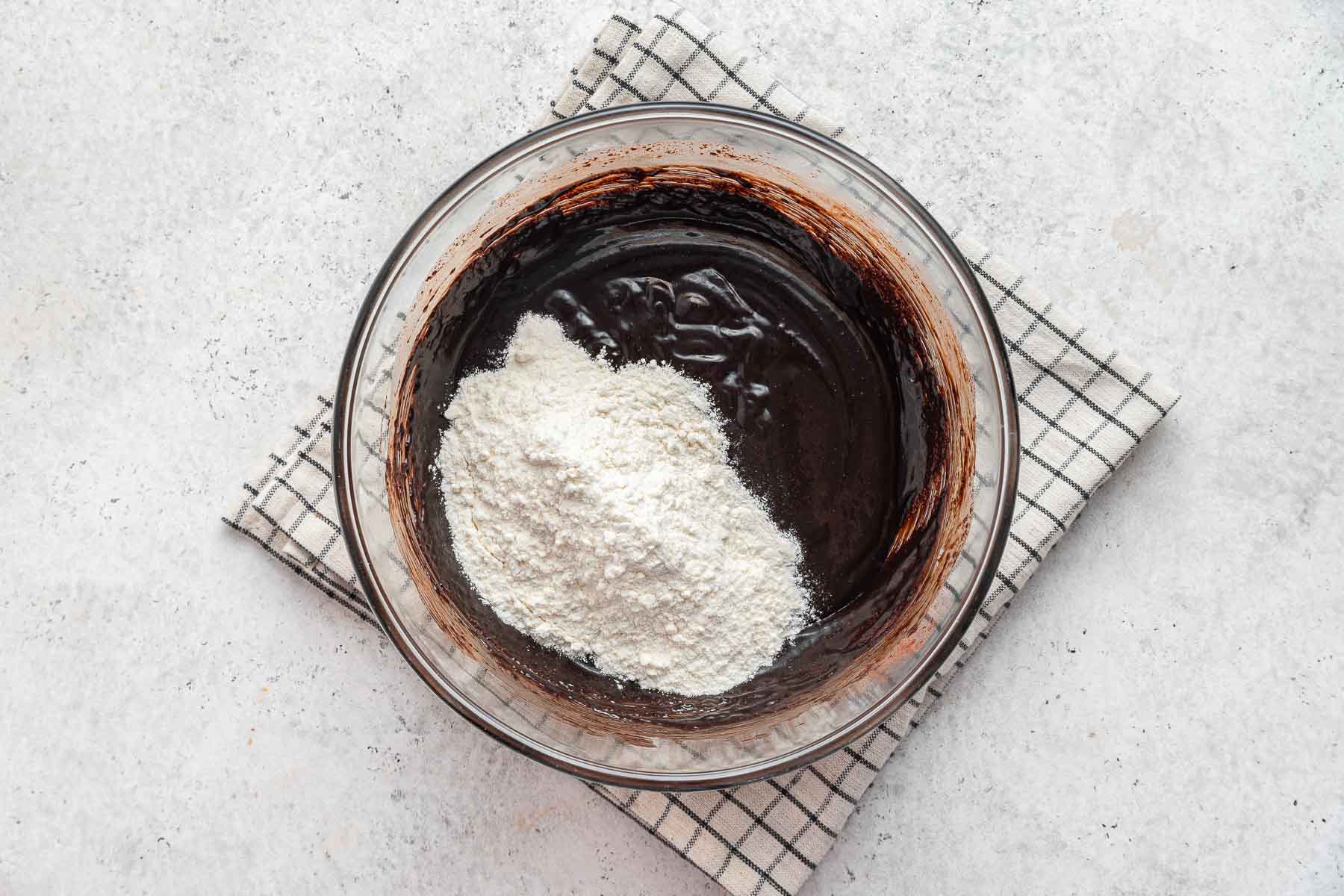 Adding flour to a raw brownie batter in a clear glass bowl.