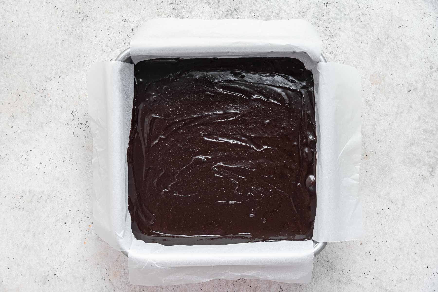 Dark brown brownie batter spread in an 8 inch square pan lined with parchment paper.