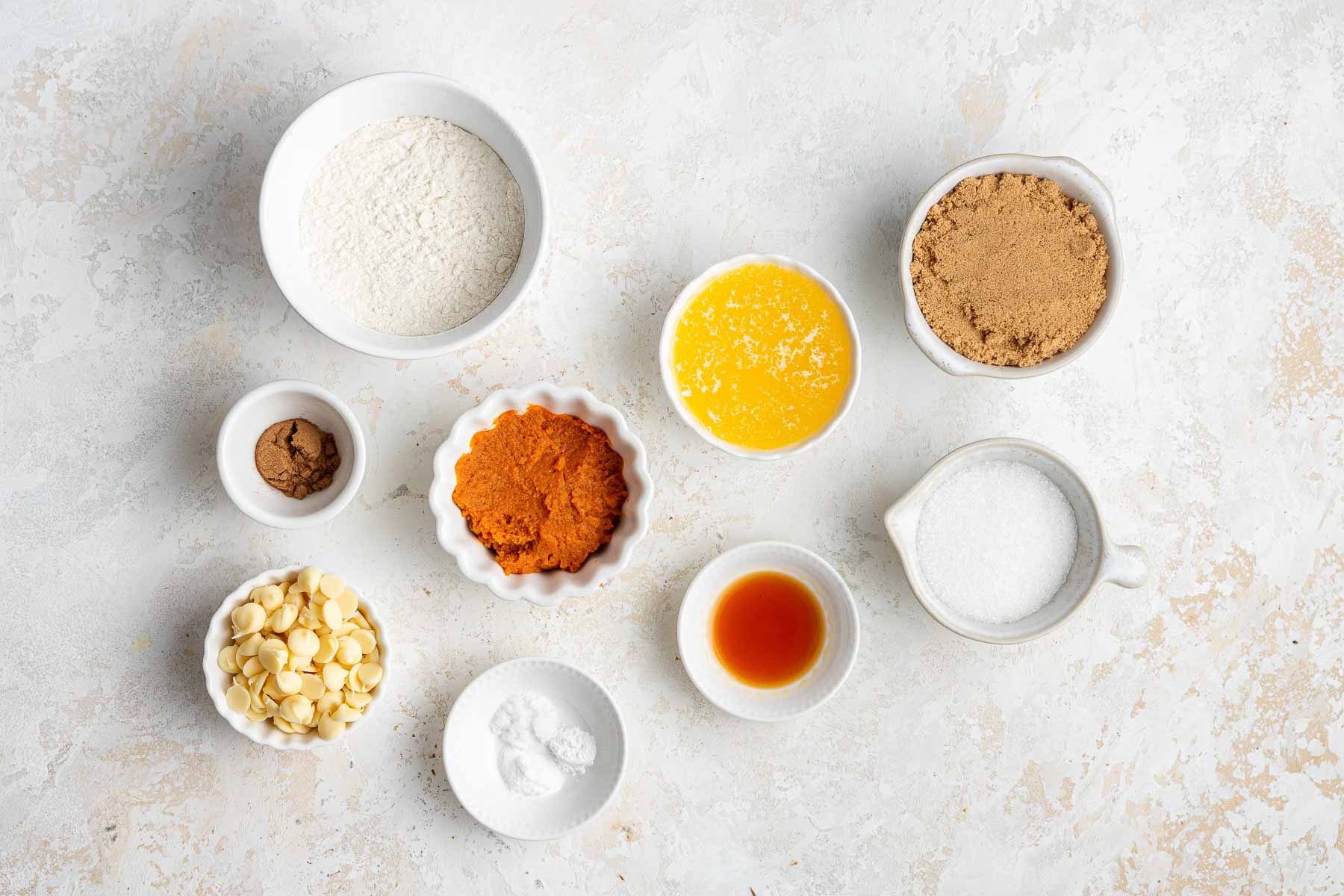 FLour, sugar, melted butter, pumpkin and spices in small bowls on white counter.