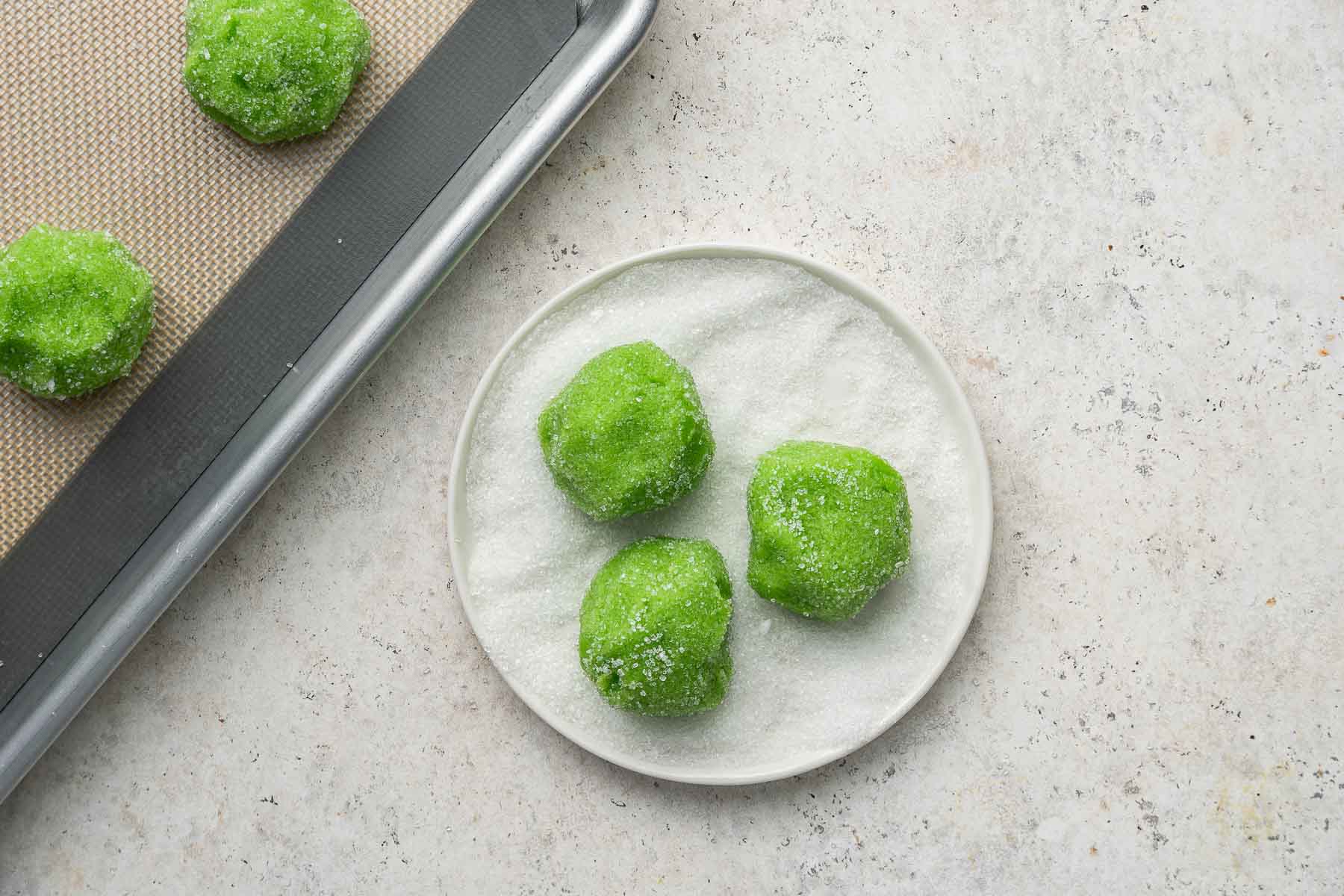 Three green dough balls being rolled in granulated sugar in small bowl.