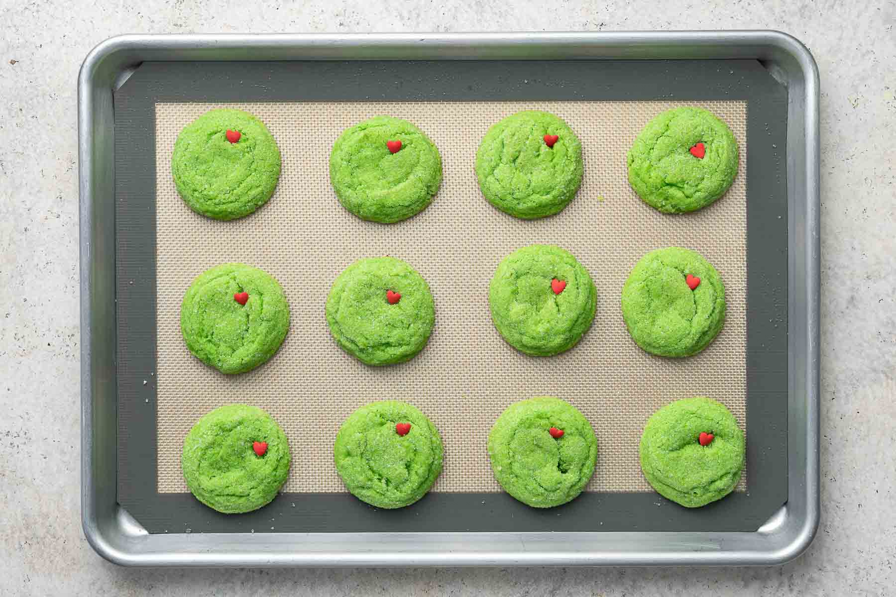 Pressing red heart shaped sprinkles into grinch sugar cookies after baking on baking sheet.