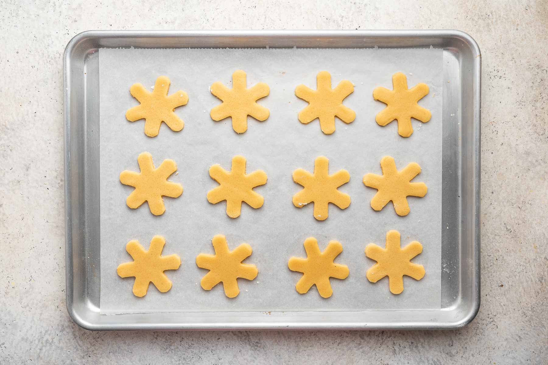 Twelve snowflake cookies on a baking sheet before being baked.