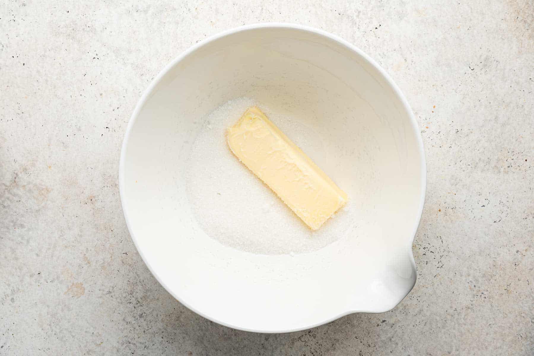 One stick of butter on top of granulated sugar in a white bowl.