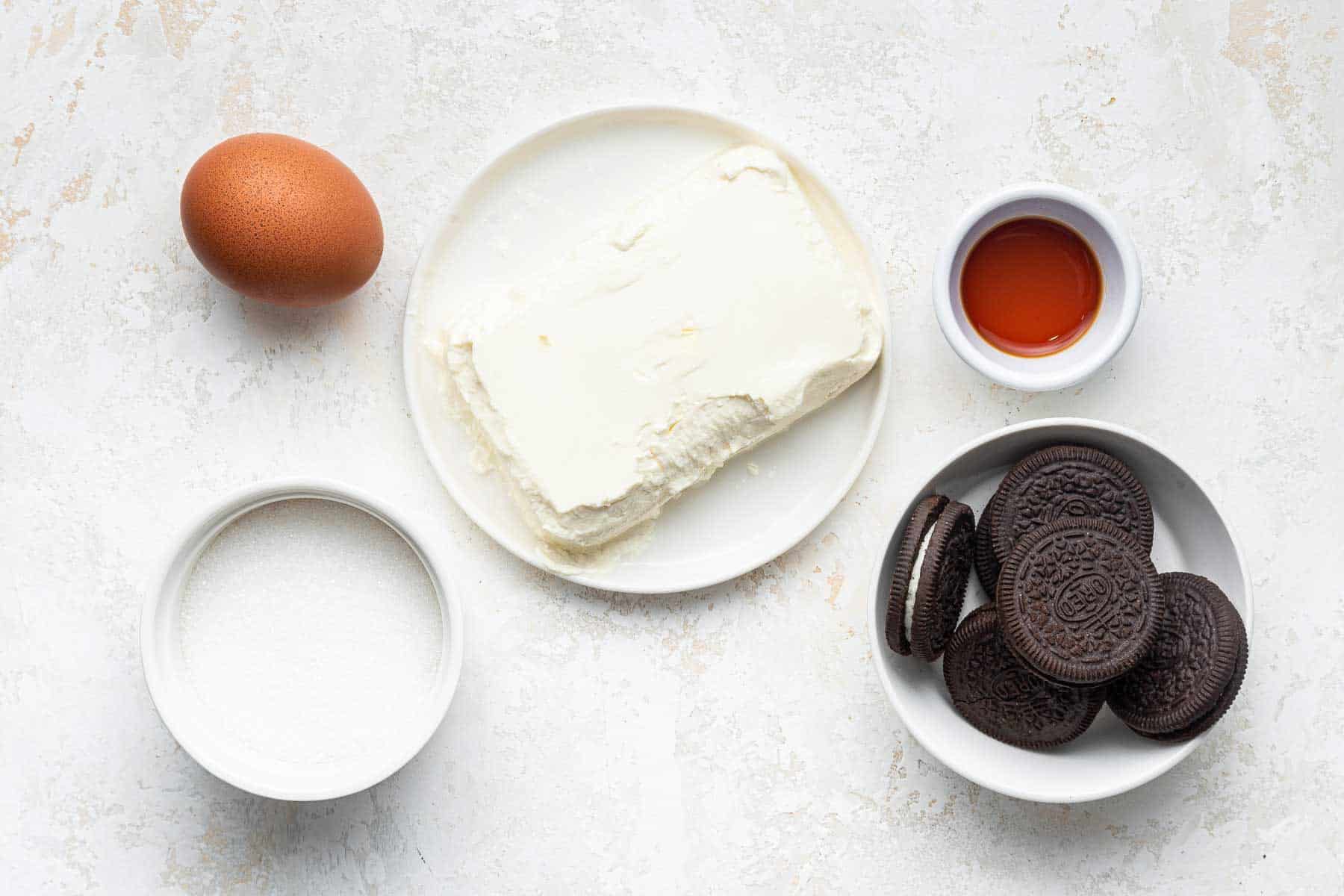 Cream cheese, egg, oreos, vanilla and sugar in small bowls on white counter.