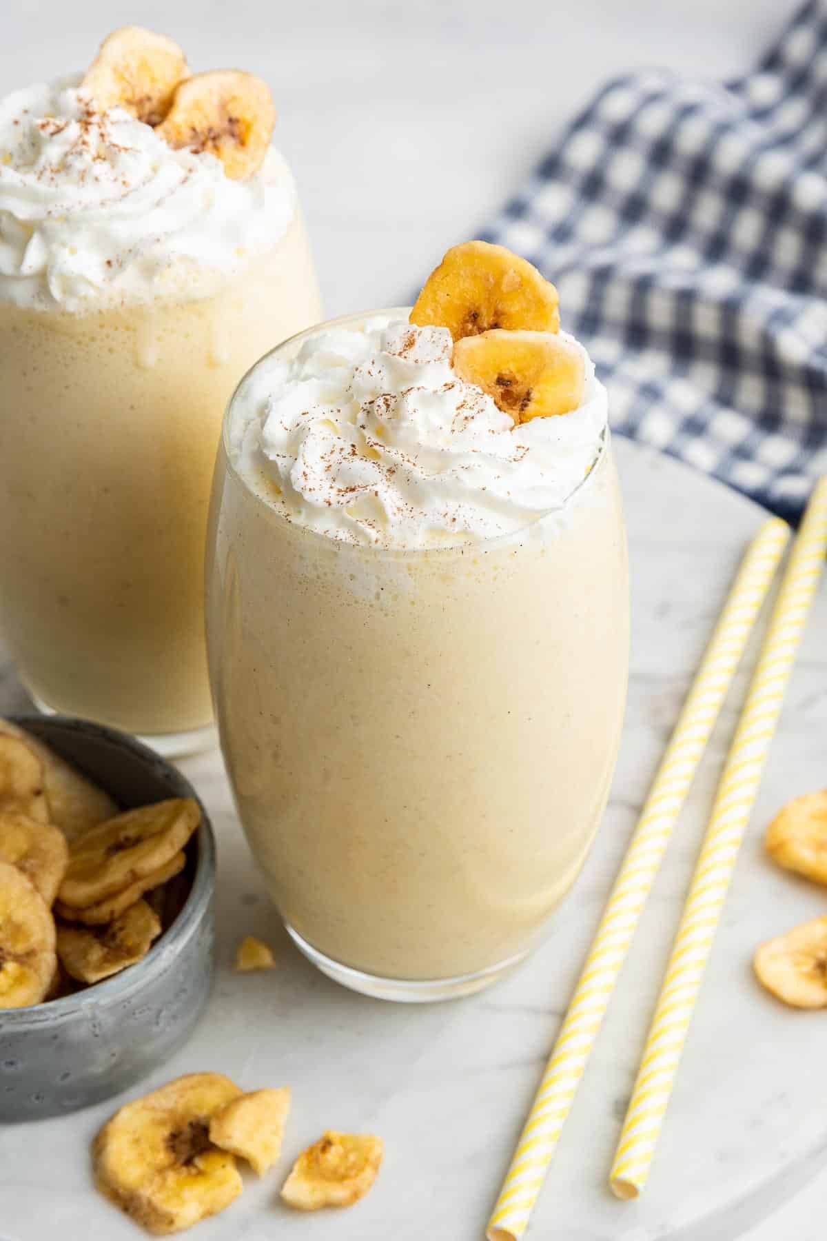 Two glasses of banana milkshake garnished with whipped cream and dried banana slices.