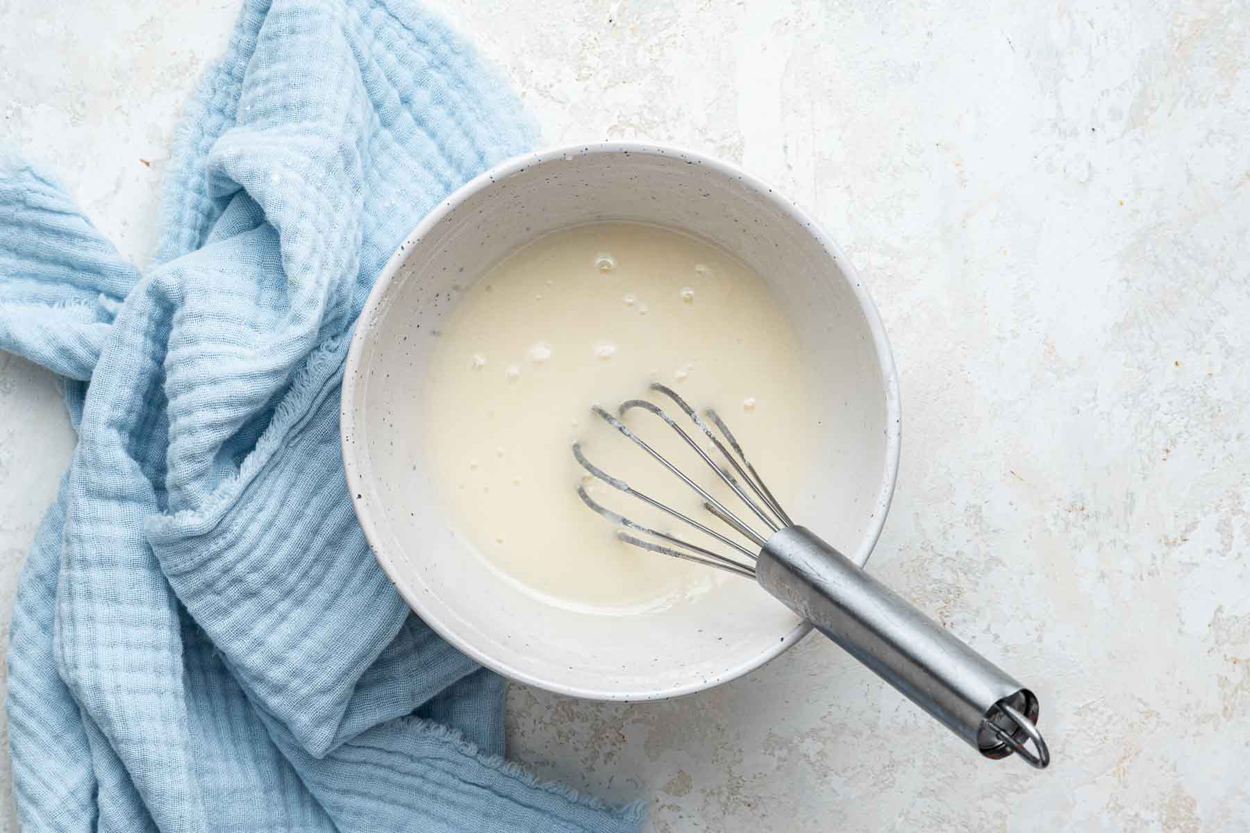 Lemon glaze in white bowl with whisk and blue kitchen towel on side.